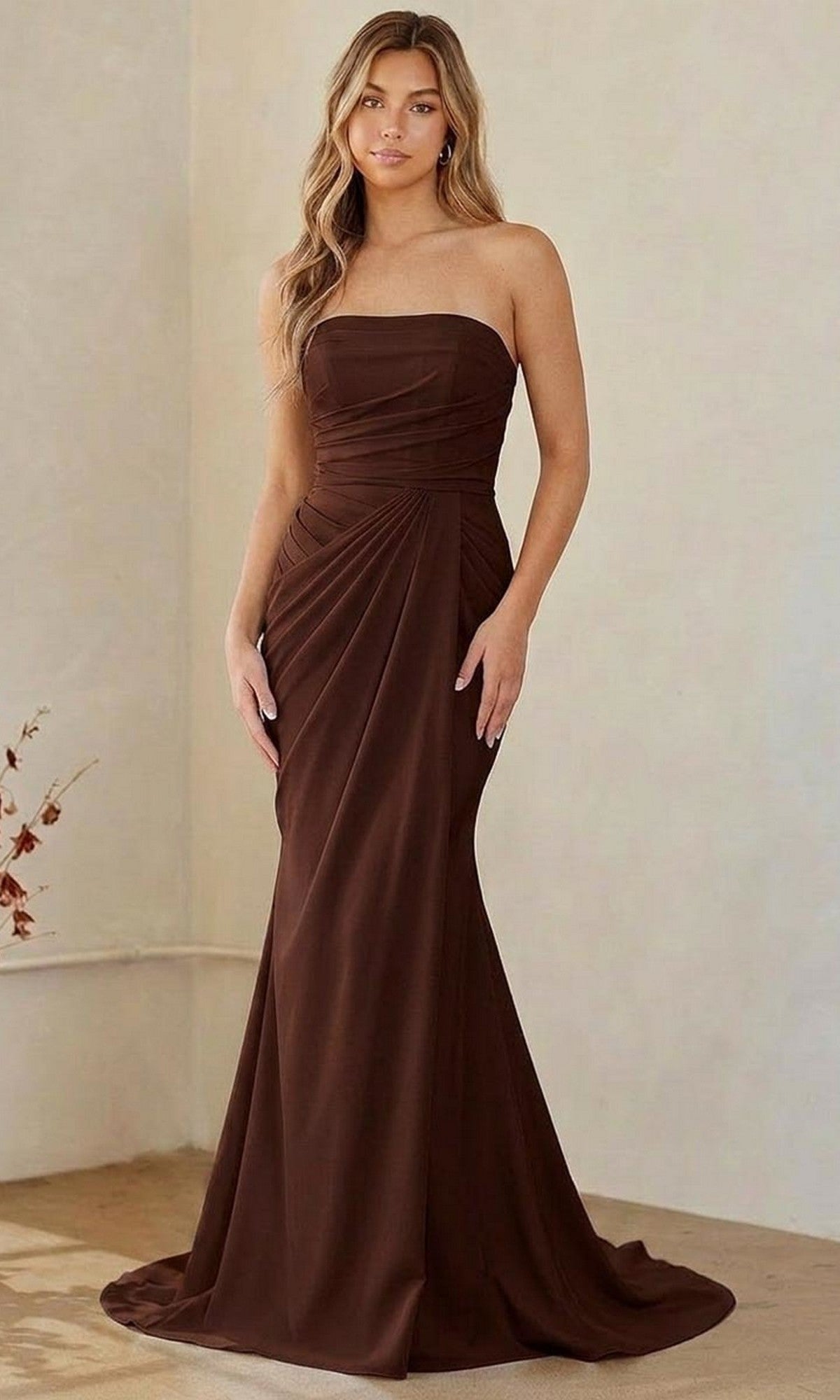 A woman in a brown Long Formal Dress: Amelia Couture 6149 with strapless, draped detailing stands indoors against a plain background.